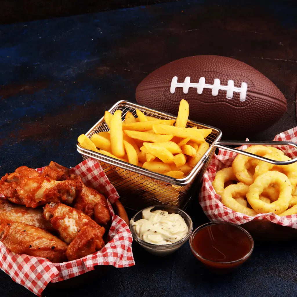 An assortment of snacks for Super Bowl Sunday. Consists of Buffalo wings, onion rings, fries, ketchup, ranch dressing in front of a football.